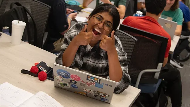 微笑的女学生举着两个大拇指，坐在桌子前拿着笔记本电脑