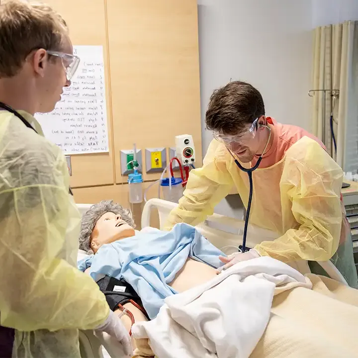 Two people in medical gowns working on a mannequin